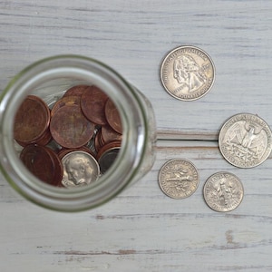 Pode incluir: Um frasco de vidro transparente cheio de moedas, incluindo centavos, com outras moedas espalhadas ao redor. As moedas estão sobre uma superfície de madeira clara.