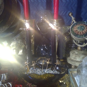 May include: Two red candles are lit and placed in front of a black, ornate candle holder. The candles are on a table with a bowl of clear glass beads and other decorative items.