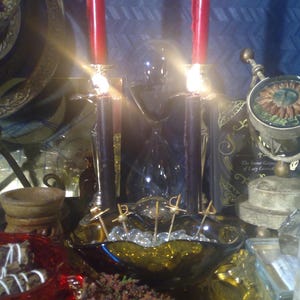 K&ouml;nnte beinhalten: Zwei rote Kerzen brennen auf einem schwarzen Metall-Kandelaber mit goldenen Akzenten. Die Kerzen stehen vor einem dunkelblauen Hintergrund mit einem wei&szlig;en Chevron-Muster. Auf dem Tisch befinden sich weitere Dekorationsartikel, darunter eine Glasschale mit klaren Perlen, eine rote Schale mit Schokoladenbonbons und ein Buch mit dem Titel "The Secret Grimoire of Lucy Cavendish".