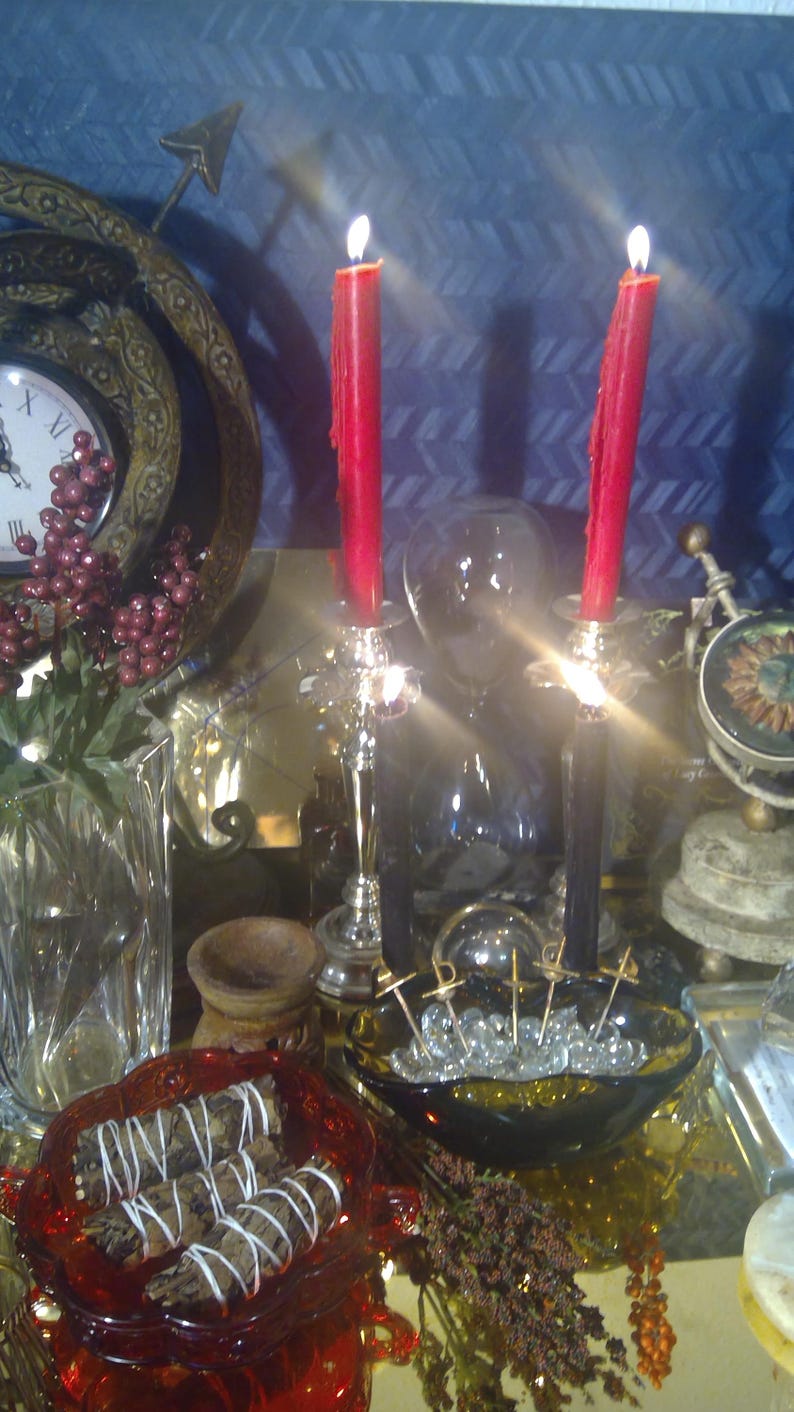K&ouml;nnte beinhalten: Eine rote Kerze brennt auf einem silbernen Kerzenleuchter, eine zweite rote Kerze brennt auf einem schwarzen Kerzenleuchter, eine klare Glaskugel, eine Glasschale gef&uuml;llt mit klaren Glasperlen und goldenen Metallnadeln, eine rote Glasschale mit drei wei&szlig;en Salbeist&auml;bchen, eine Holzschale, eine klare Glasvase mit einer gr&uuml;nen Pflanze und roten Beeren und eine goldene Metalluhr mit r&ouml;mischen Ziffern.