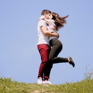 Könnte beinhalten: Ein Paar umarmt sich und küsst sich auf einer grünen Wiese. Der Mann trägt ein weißes Hemd und eine rote Hose. Die Frau trägt ein Blusenkleid, eine schwarze Hose und schwarze Schuhe. Der Himmel ist blau.