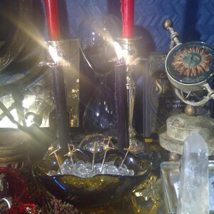 May include: A black glass bowl filled with clear glass beads and topped with silver pins. Two red candles are lit in gold candle holders. A clear glass hourglass is in the background.