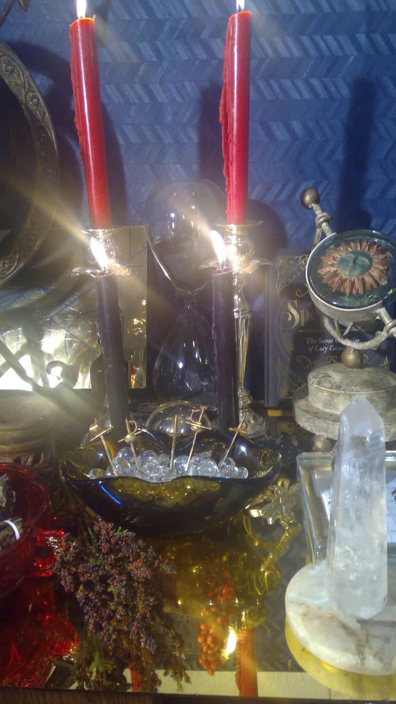 May include: A black glass bowl filled with clear glass beads and topped with silver pins. Two red candles are lit in gold candle holders. A clear glass hourglass is in the background.