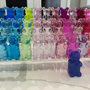 May include: A collection of gummy bear shaped resin figurines in various colors, including blue, green, pink, purple, and clear. The figurines are arranged on a clear acrylic stand.