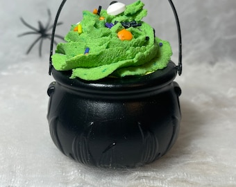 May include: A black cauldron-shaped container with a handle, filled with green frosting and topped with colorful sprinkles and a candy eye. The cauldron has decorative vertical lines and is set against a soft, white background. A black spider decoration is in the background.