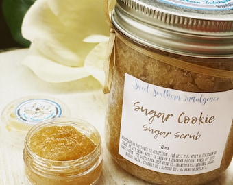Może przedstawiać: Szklany słoik z cukrowym peelingiem o smaku ciasteczek z cukrem, z brązową etykietą z napisem "Sweet Southern Indulgence Sugar Cookie Sugar Scrub 8 oz". Słoik jest przewiązany brązową wstążką i ma srebrną pokrywkę z białą naklejką przedstawiającą kwiat magnolii.