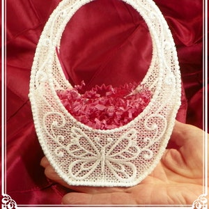 May include: White lace oval-shaped basket with a butterfly design and a pink ribbon handle. The basket is filled with pink tissue paper.