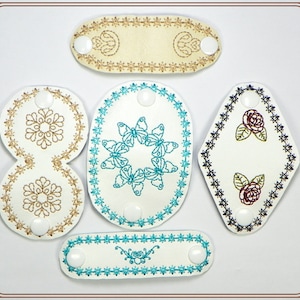 May include: Five white leather patches with different embroidered designs. The designs include a butterfly, a rose, and a snowflake. The patches have a white background and are edged with a blue or brown stitching.