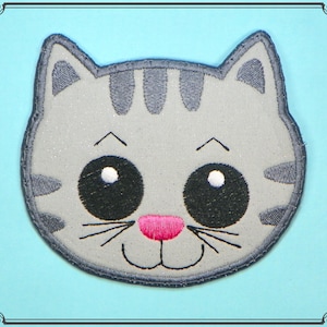 May include: A gray and white embroidered cat face with black eyes and a pink nose. The cat has a happy expression and is smiling.