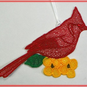 Könnte beinhalten: Roter Kardinal-Anhänger mit einem grünen Blatt und gelben Blumen. Der Anhänger ist aus Spitze gefertigt und hat ein weißes Band zum Aufhängen.