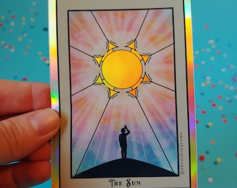 Puede incluir: Una carta de tarot holográfica con un sol amarillo y una silueta de una persona levantando la mano en el aire. La carta se titula "El Sol" y tiene un fondo de acuarela rosa claro y azul.