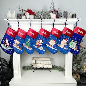 Puede incluir: Siete calcetines de Navidad de terciopelo azul con puños de fieltro rojo, personalizados con nombres y con escenas de invierno bordadas. Los calcetines están colgados en una repisa de chimenea blanca sobre una chimenea.
