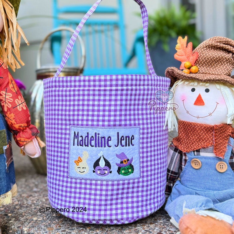 May include: A purple and white gingham Halloween trick-or-treat bag with a handle. The bag has a rectangular patch with the name "Madeline Jene" and three embroidered Halloween character faces.
