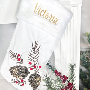 May include: White Christmas stocking with a faux fur cuff, embroidered with the name "Victoria". The stocking features a gold and brown sequined pine cone design with red berries.