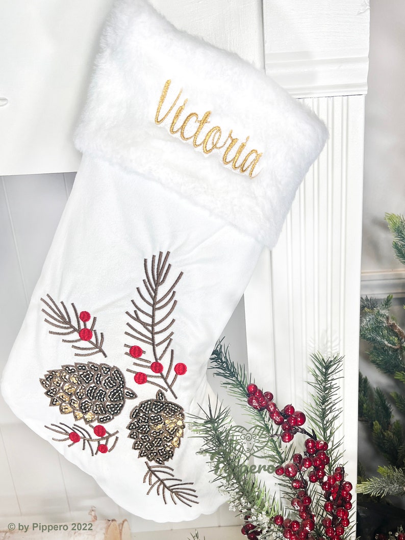 May include: White Christmas stocking with a faux fur cuff, embroidered with the name "Victoria". The stocking features a gold and brown sequined pine cone design with red berries.