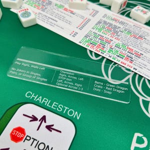 以下が含まれることがあります： 緑色の麻雀ゲームボード、白い牌、透明なプラスチックオーバーレイ。 オーバーレイには「Charleston」や「STOP-tion」など、ゲームプレイを説明するテキストがあります。 追加のゲーム手順も表示されています。