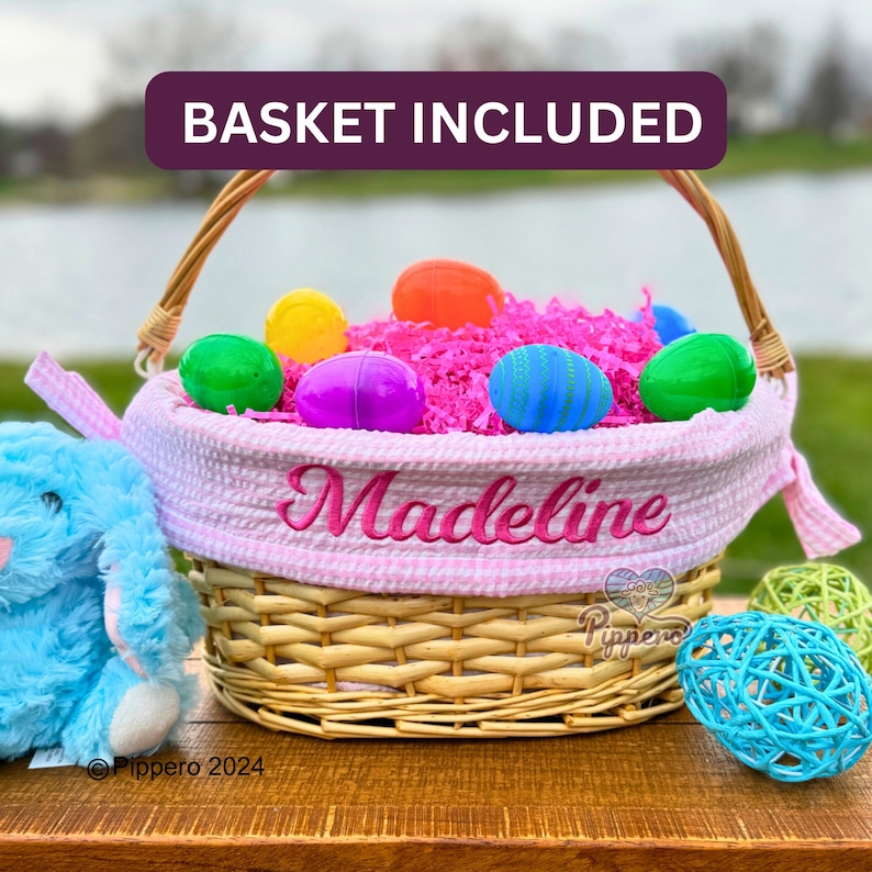 Pode incluir: Uma cesta de P&aacute;scoa de vime com forro rosa xadrez e o nome "Madeline" bordado em rosa. A cesta &eacute; preenchida com papel rosa e ovos de pl&aacute;stico coloridos. Um coelho de pel&uacute;cia azul est&aacute; ao lado da cesta.