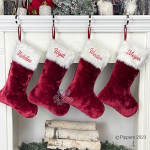 May include: Four red velvet Christmas stockings with white faux fur trim and embroidered names: Madeline, Wyatt, Winston, and Megan. Each stocking hangs from a silver hook with a red ribbon.