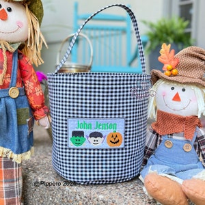 May include: Black and white gingham Halloween bucket with a handle. The bucket has a rectangular patch with the name "John Jenson" and three embroidered Halloween characters: a green monster, a vampire, and a jack-o'-lantern.