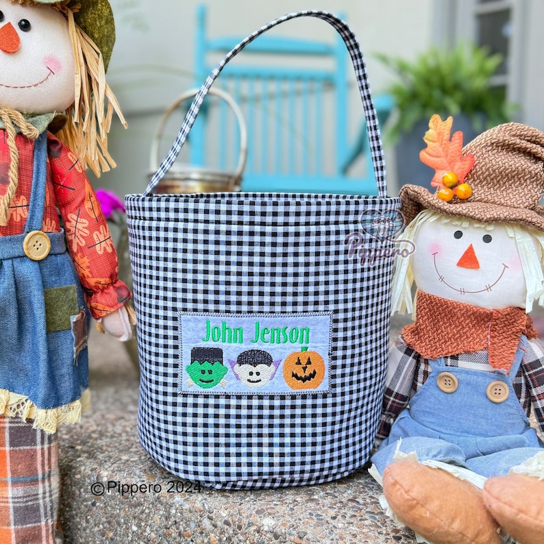 May include: Black and white gingham Halloween bucket with a handle. The bucket has a rectangular patch with the name "John Jenson" and three embroidered Halloween characters: a green monster, a vampire, and a jack-o'-lantern.