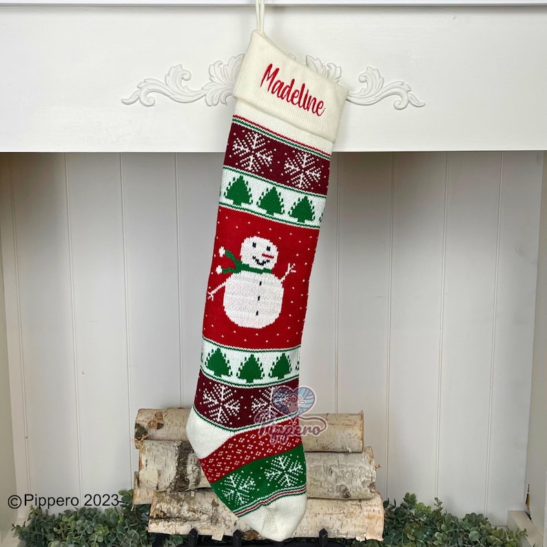 May include: White knitted Christmas stocking with a red, green, and white snowman design. The stocking has the name "Madeline" embroidered on the cuff.