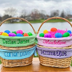 May include: Two woven Easter baskets with handles, filled with colorful eggs and decorative paper. The baskets have gingham fabric linings and personalized names embroidered on them. One basket is green, the other is pink, and the bottom baskets are blue and purple.