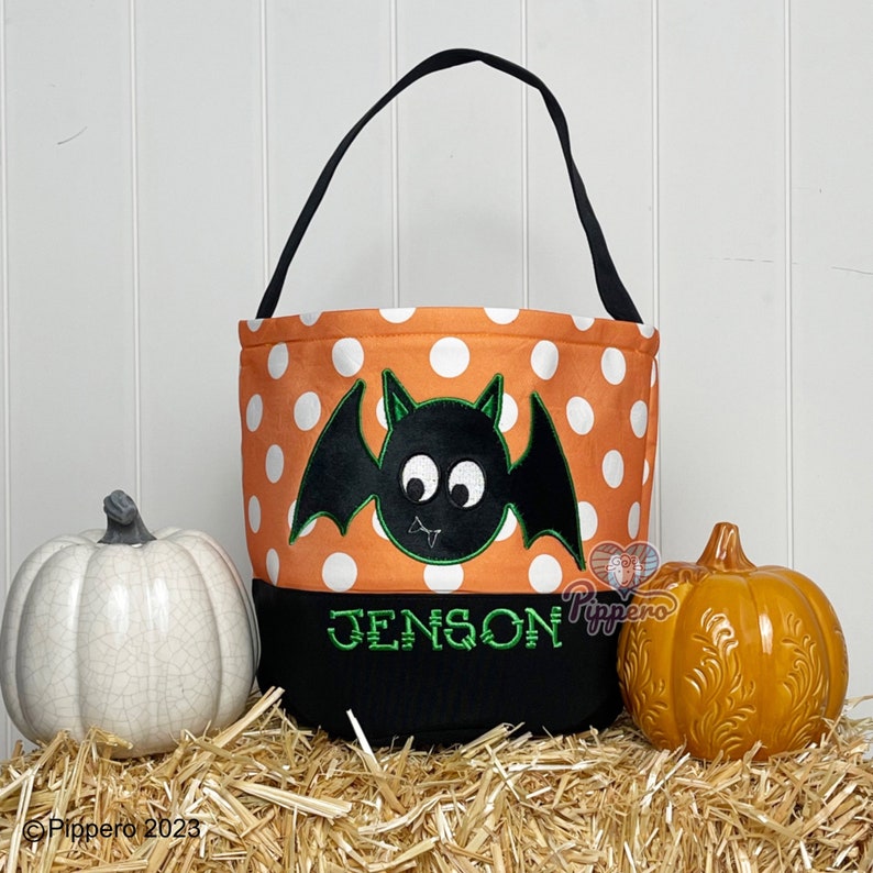 May include: Orange and white polka dot trick or treat bag with a black bat with green eyes and wings. The bag has a black bottom with the name "Jenson" embroidered in green.
