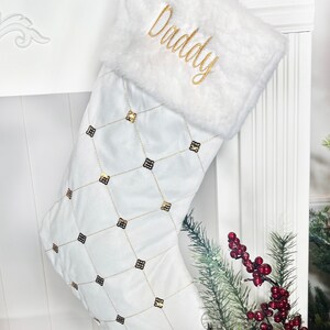 May include: White Christmas stocking with a gold and black diamond pattern and a fluffy white cuff. The stocking is personalized with the word "Daddy" in gold embroidery.