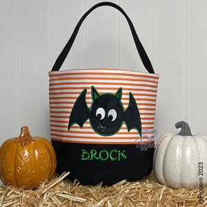 May include: Orange and black striped Halloween trick or treat bag with a black bat with green eyes and a green mouth. The bag has a black bottom with the name Brock embroidered in green.