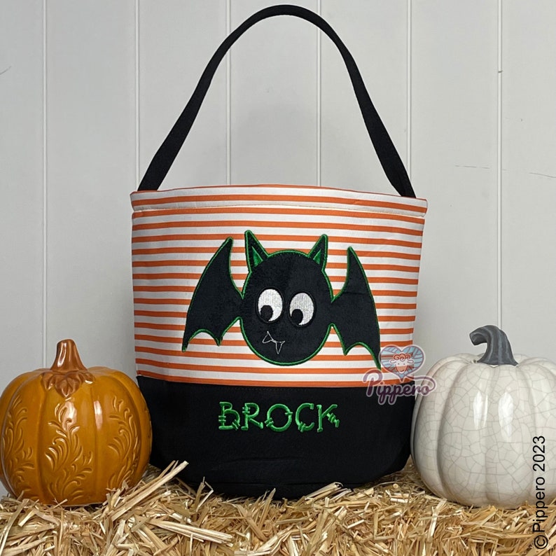 May include: Orange and black striped Halloween trick or treat bag with a black bat with green eyes and a green mouth. The bag has a black bottom with the name Brock embroidered in green.