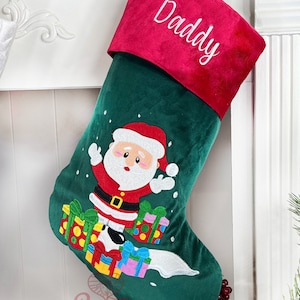 May include: Green Christmas stocking with a red cuff and the word "Daddy" embroidered on it. Santa Claus is embroidered on the stocking with presents and snowflakes.