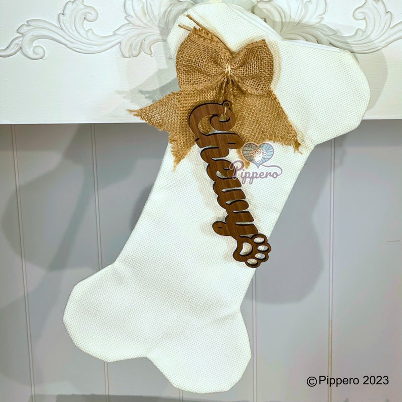 May include: White dog bone-shaped Christmas stocking with a burlap bow and a wooden name tag that says "Chewy" with a paw print.
