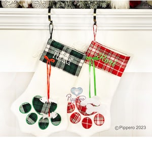 May include: Two personalized Christmas stockings with paw print designs. The stocking on the left is green and white plaid with the name "Tucker" embroidered on it. The stocking on the right is red and white plaid with the name "Rori" embroidered on it.