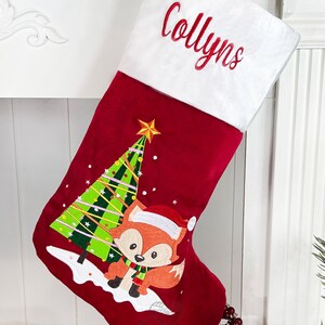 May include: Red velvet Christmas stocking with a white cuff, personalized with the name "Collyn's". The stocking features an embroidered design of a fox standing in front of a Christmas tree.