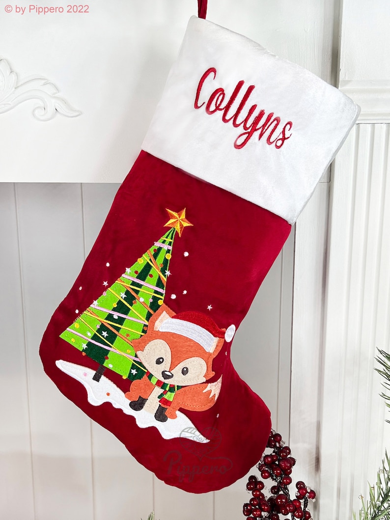 May include: Red velvet Christmas stocking with a white cuff, personalized with the name "Collyn's". The stocking features an embroidered design of a fox standing in front of a Christmas tree.