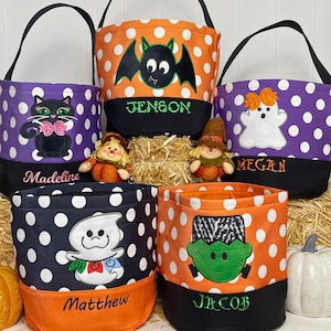 May include: Five fabric Halloween buckets with handles, featuring polka dot patterns in purple, orange, and black. Each bucket is embroidered with a character: a black cat, a bat, a ghost, a second ghost, and Frankenstein, plus names Madeline, Jenson, Megan, Matthew, and Jacob.