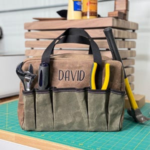 May include: A brown canvas tool bag with black handles and multiple pockets. The name "DAVID" is embroidered on the front. Tools visible include a wrench, screwdriver, pliers, and a hammer. The bag is on a green cutting mat.