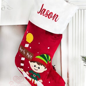 May include: Red Christmas stocking with a white cuff, personalized with the name "Jason". The stocking features an embroidered elf with a green hat and a sign that reads "North Pole".