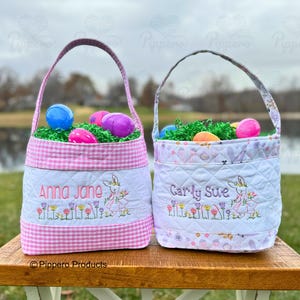 May include: Two pink and white Easter baskets with gingham trim. The baskets are filled with green grass and plastic Easter eggs. One basket is embroidered with the name "Anna Jane" and a bunny holding a basket of eggs. The other basket is embroidered with the name "Carly Sue" and a bunny holding a basket of eggs.