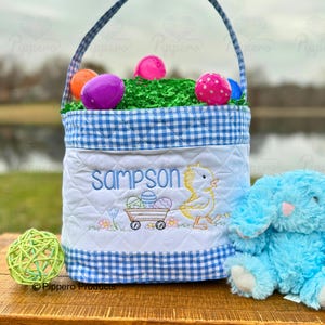 May include: Blue and white gingham Easter basket with a personalized name "Sampson" embroidered on the front. The basket is filled with colorful plastic Easter eggs and green grass. A small yellow chick with a wagon full of eggs is embroidered below the name. A blue plush bunny sits next to the basket.