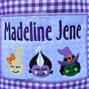May include: Embroidered patch with the name "Madeline Jene" in purple thread. Below the name are three cartoon Halloween characters: a ghost with an orange bow, a purple monster, and a green witch with a purple hat. The background is light blue.