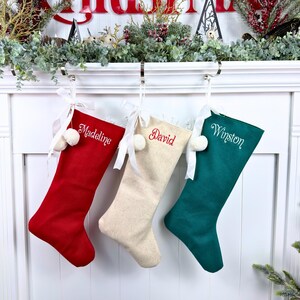 May include: Three Christmas stockings in red, cream, and dark green fabric. Each stocking features a white ribbon bow, two small white pom-poms, and embroidered names: Madeline on the red, David on the cream, and Winston on the green.
