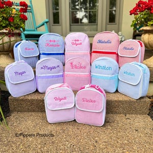 May include: A collection of personalized backpacks and lunch bags in various colors, including pink, blue, and purple. Each bag features a name embroidered in a coordinating color. The bags have a striped pattern and are arranged on a stone surface.