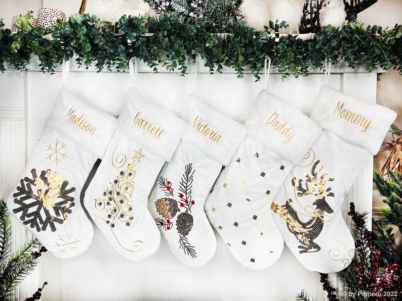 May include: Five white Christmas stockings with gold embroidery and embellishments. The stockings are personalized with the names "Madison", "Barrett", "Victoria", "Daddy", and "Mommy".