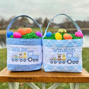 May include: Two blue and white gingham Easter baskets filled with colorful eggs and green filler. Each basket has a name embroidered on the front, along with a train design featuring a bunny and chick.