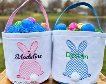 May include: Two personalized Easter baskets with bunny designs. The blue basket has the name "Declan" embroidered on it and the pink basket has the name "Madeline" embroidered on it. Both baskets are filled with colorful plastic Easter eggs and blue shredded paper.