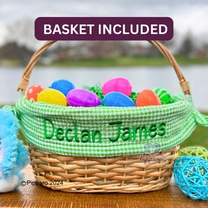 Pode incluir: Uma cesta de P&aacute;scoa de vime tran&ccedil;ado cheia de ovos de pl&aacute;stico coloridos e grama decorativa verde. A cesta tem um forro xadrez verde e branco com o nome "Declan James" bordado em verde. O texto "BASKET INCLUDED" est&aacute; no topo.