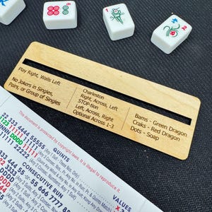 May include: A wooden Mahjong cheat sheet with text, alongside white Mahjong tiles. The cheat sheet has text including "Charleston" and "Bams - Green Dragon". The tiles have red and green markings. A printed document with text is also visible.