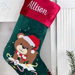 May include: Green velvet Christmas stocking with a red felt cuff. The cuff is embroidered with the name "Allison". The stocking features an embroidered brown bear wearing a red Santa hat and a white scarf. The bear is surrounded by candy canes and snowflakes.