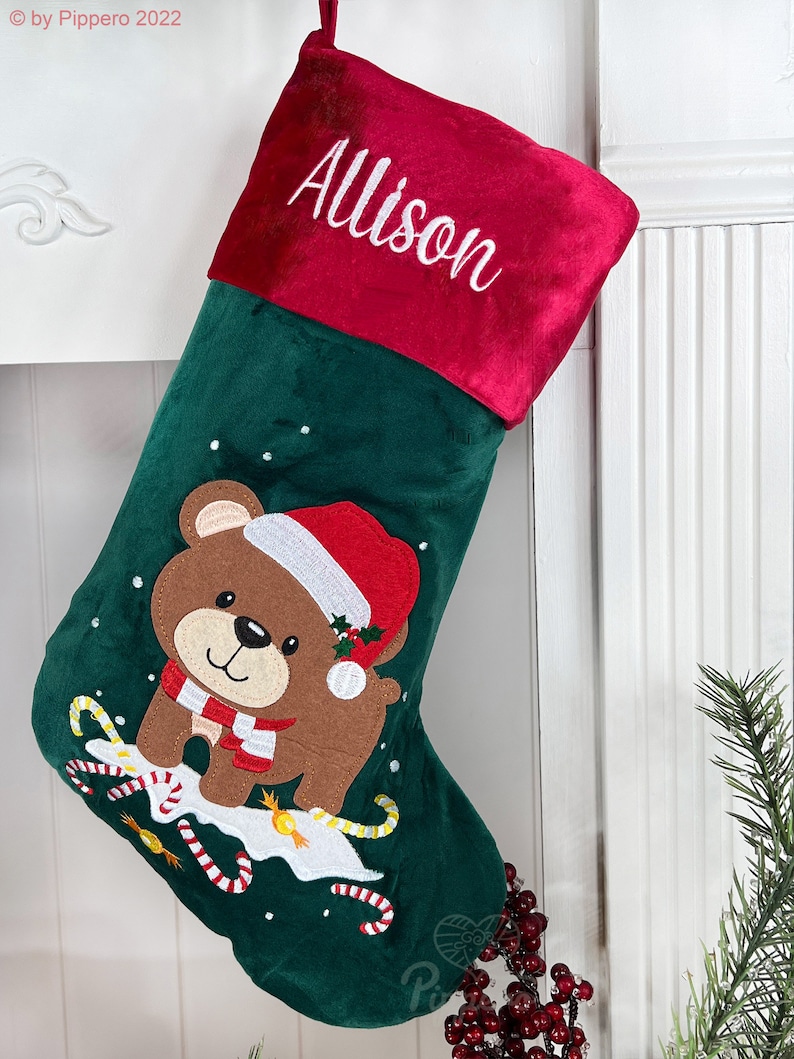 May include: Green velvet Christmas stocking with a red felt cuff. The cuff is embroidered with the name "Allison". The stocking features an embroidered brown bear wearing a red Santa hat and a white scarf. The bear is surrounded by candy canes and snowflakes.